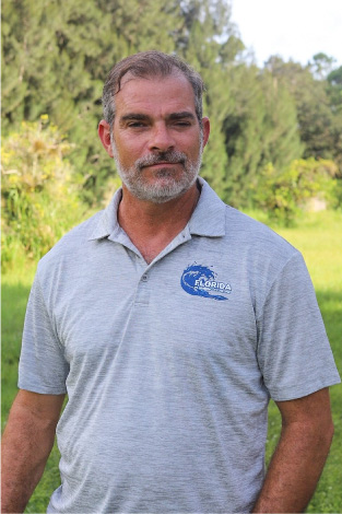 David, Owner Operator of Florida Plumbing Plus, wearing a branded gray polo shirt, standing outdoors in a lush green setting.